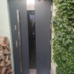Nowoczesne, grafitowe drzwi zewnętrzne z długim, pionowym uchwytem i przeszklonym panelem, obok ściana porośnięta bluszczem, Niepołomice.