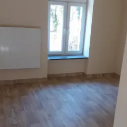 Jasne wnętrze pokoju po remoncie, z panelami podłogowymi imitującymi drewno, grzejnikiem na ścianie i oknem z widokiem na drzewa.