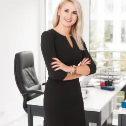 Portret uśmiechniętej blondynki w czarnej sukience stojącej na tle biura z biurkiem, krzesłem i oknem.