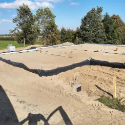 Przygotowany teren pod budowę domu jednorodzinnego z widocznymi zbrojeniami, izolacją pionową fundamentów i elementami instalacji, w tle zielony krajobraz z drzewami i zabudowaniami.