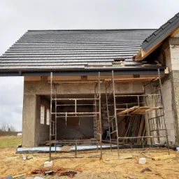 Surowy stan budynku z pustaków z zamontowaną częścią dachu z grafitowej dachówki ceramicznej, widoczne rusztowanie, teren budowy z gruzem i wystającą rurą kanalizacyjną.