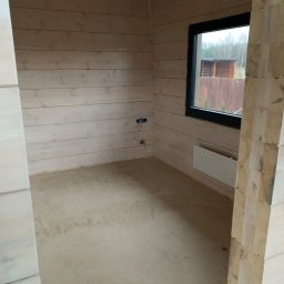 Firma Wielobranżowa Przemysław Rycharski - Surowy stan wnętrza domu z bali w Lipnie, widoczne przygotowane ściany z drewna, okno z widokiem na zewnątrz i wylewka na podłodze.