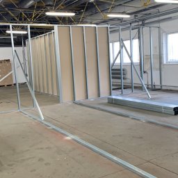 Firma Wielobranżowa Przemysław Rycharski - Montaż ścian działowych z płyt gipsowo-kartonowych na metalowym stelażu w surowym stanie, widoczne profile podłogowe z zaznaczonym położeniem, industrialne wnętrze z instalacjami pod sufitem.