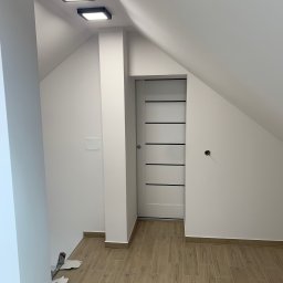 Firma Wielobranżowa Przemysław Rycharski - Poddasze po remoncie: białe ściany, drewnopodobna podłoga z płytek, przesuwne drzwi z poziomymi czarnymi paskami, dwa kwadratowe czarne plafony LED, widoczne niedokończone gniazdko elektryczne...