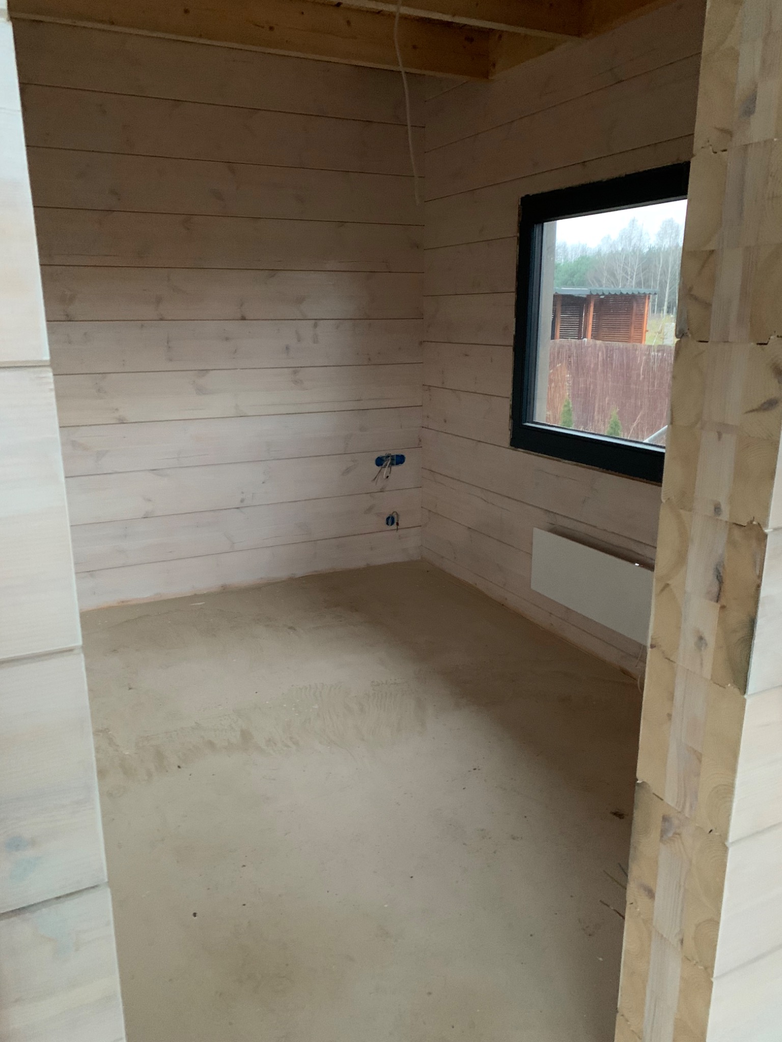Surowy stan wnętrza domu z bali w Lipnie, widoczne przygotowane ściany z drewna, okno z widokiem na zewnątrz i wylewka na podłodze.