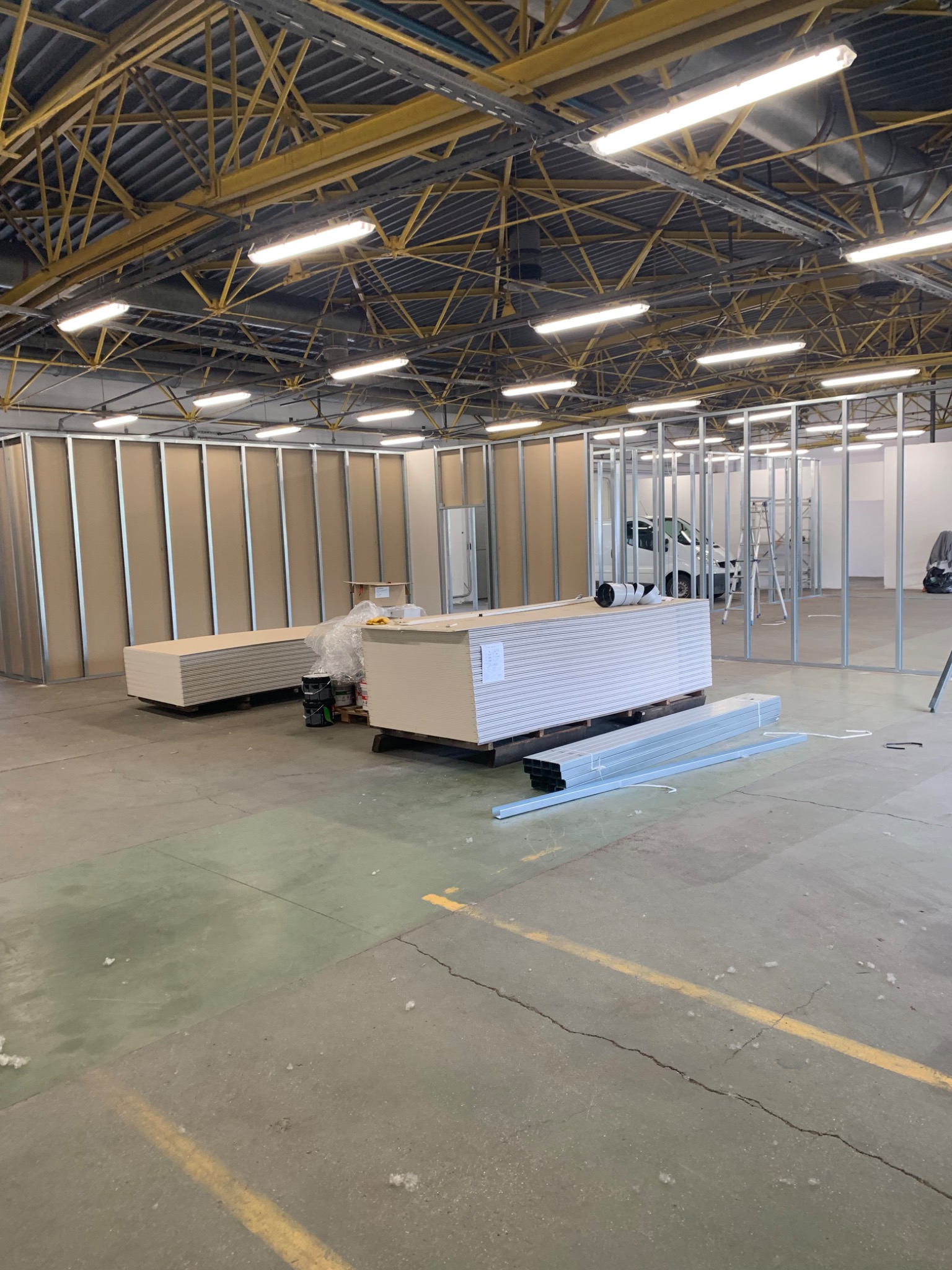 Montaż ścian działowych z płyt gipsowo-kartonowych w hali przemysłowej, widoczne profile metalowe, ułożone płyty, drabina i dostawczy samochód w tle.