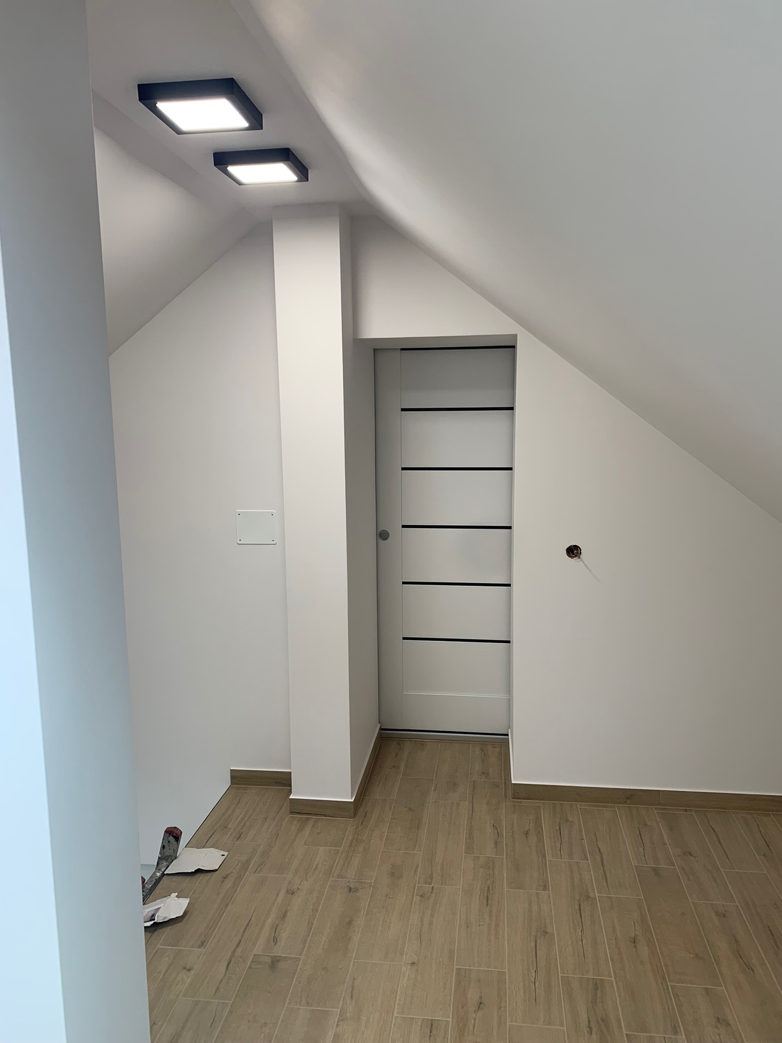 Poddasze po remoncie: białe ściany, drewnopodobna podłoga z płytek, przesuwne drzwi z poziomymi czarnymi paskami, dwa kwadratowe czarne plafony LED, widoczne niedokończone gniazdko elektryczne...