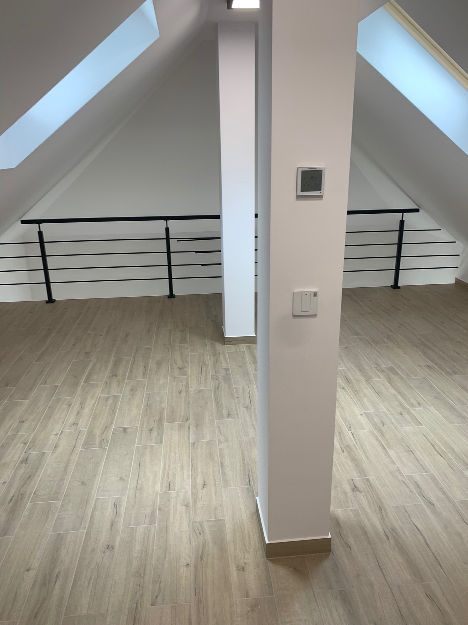 Poddasze wykończone płytkami imitującymi drewno, z widocznymi skosami, oknami dachowymi, balustradą z czarnych prętów i termostatem na białym filarze.