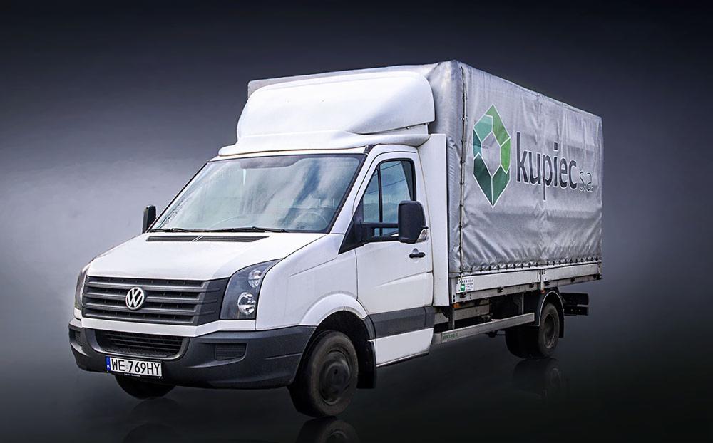 Biały samochód dostawczy Volkswagen z plandeką z logo firmy Kupiec S.A. na tle szarego gradientu.