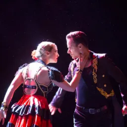 Para tancerzy w strojach scenicznych, kobieta w czerwono-czarnej sukience z falbanami, mężczyzna w czarnym fraku ze złotymi zdobieniami, w trakcie występu, oświetlenie sceniczne.