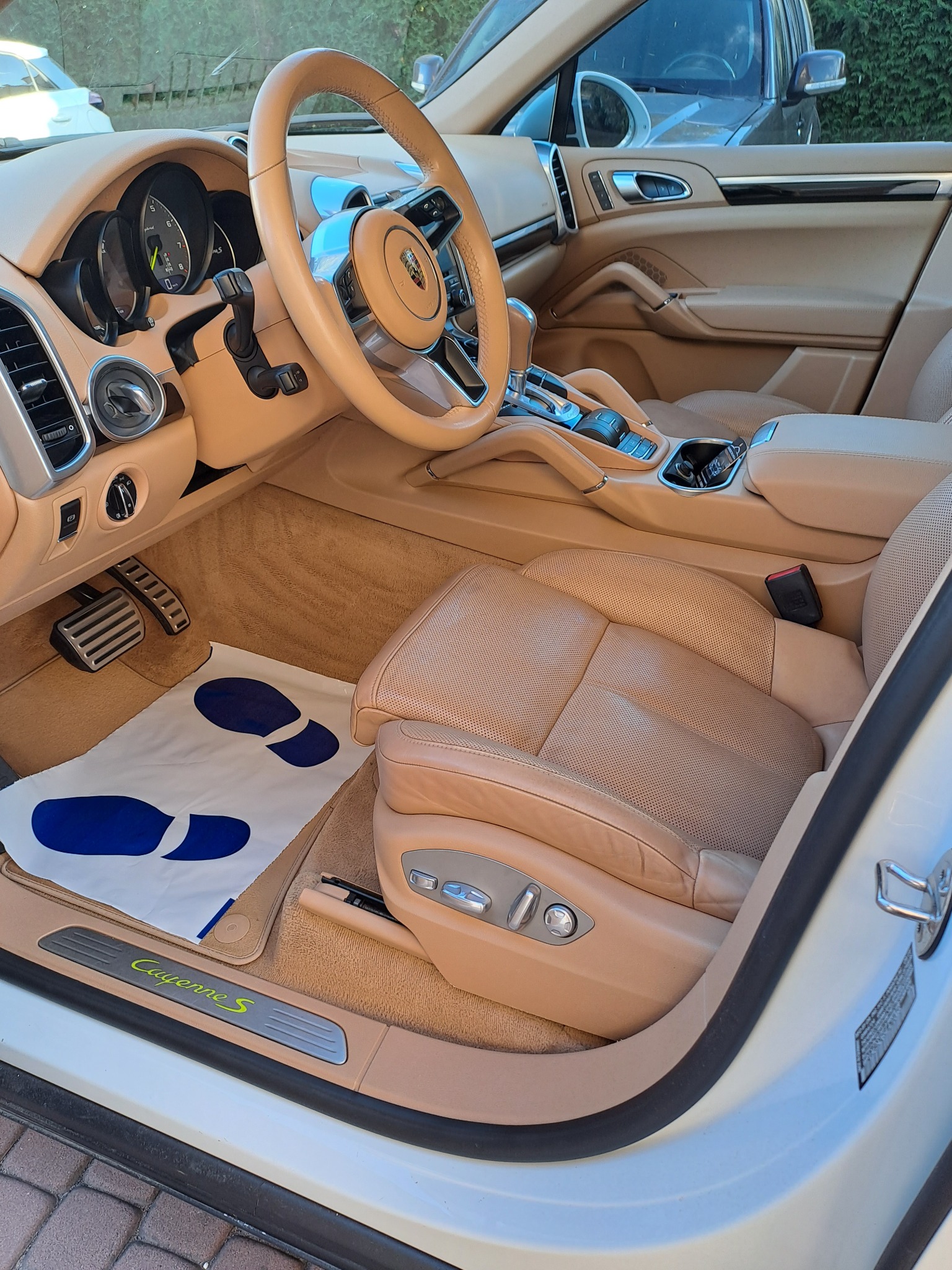 Beżowe skórzane wnętrze samochodu Porsche Cayenne S, widok na fotel kierowcy z podłożoną papierową matą ochronną na podłodze, detale deski rozdzielczej i kierownicy.