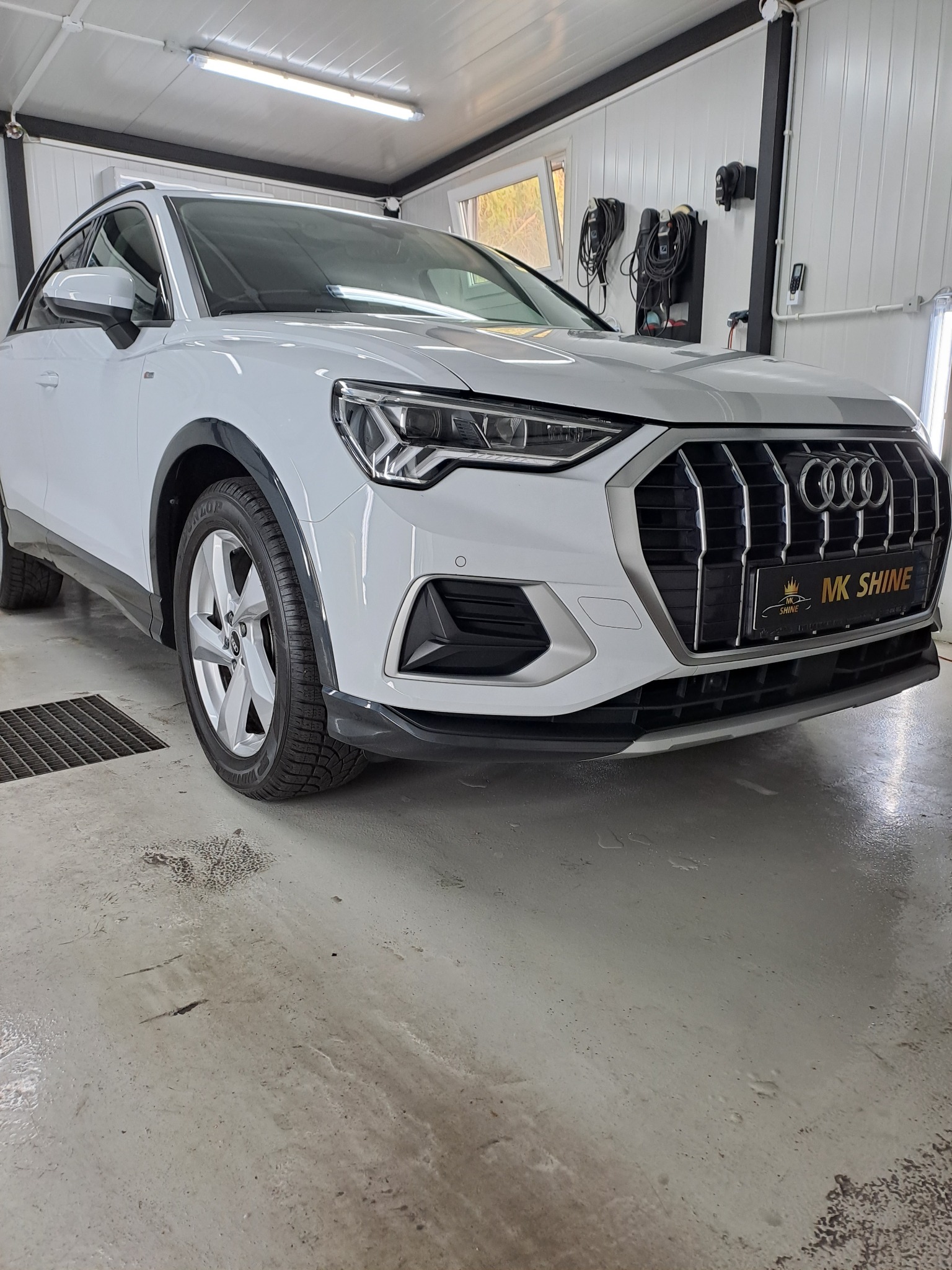 Biały samochód Audi Q3 po umyciu w myjni, widoczne ślady wody na podłodze i logo firmy 'MK Shine' na grillu.