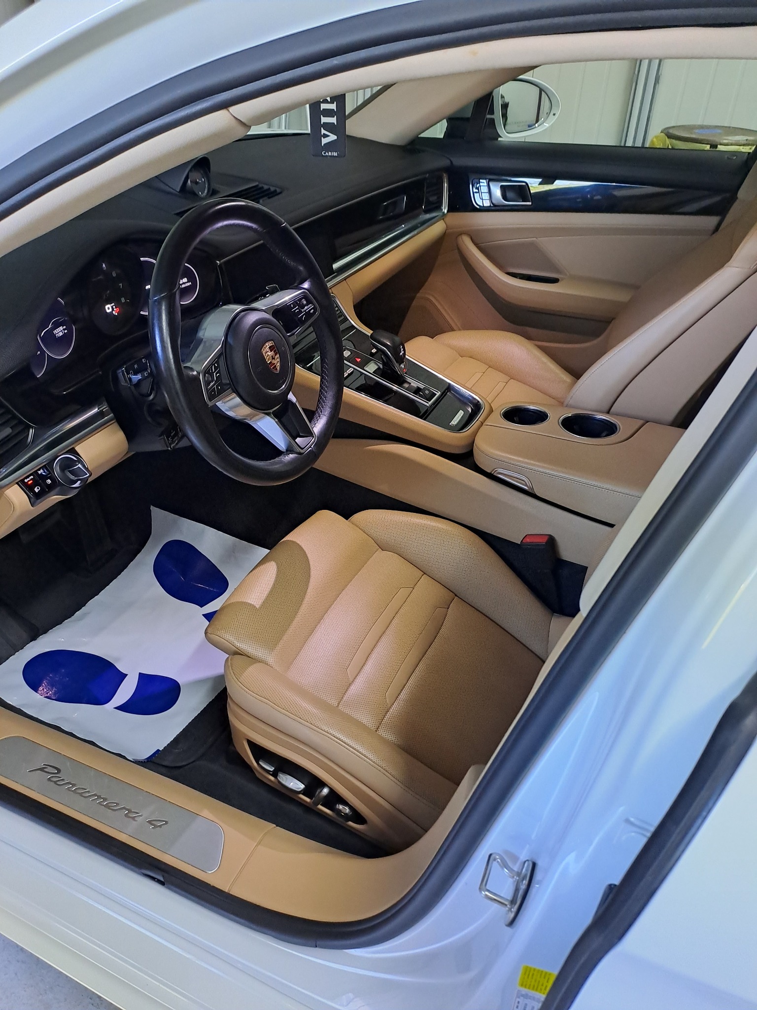 Wnętrze samochodu Porsche Panamera 4 z jasną skórzaną tapicerką, widoczne fotele, kierownica z logo Porsche oraz ochronny papierowy dywanik na podłodze.