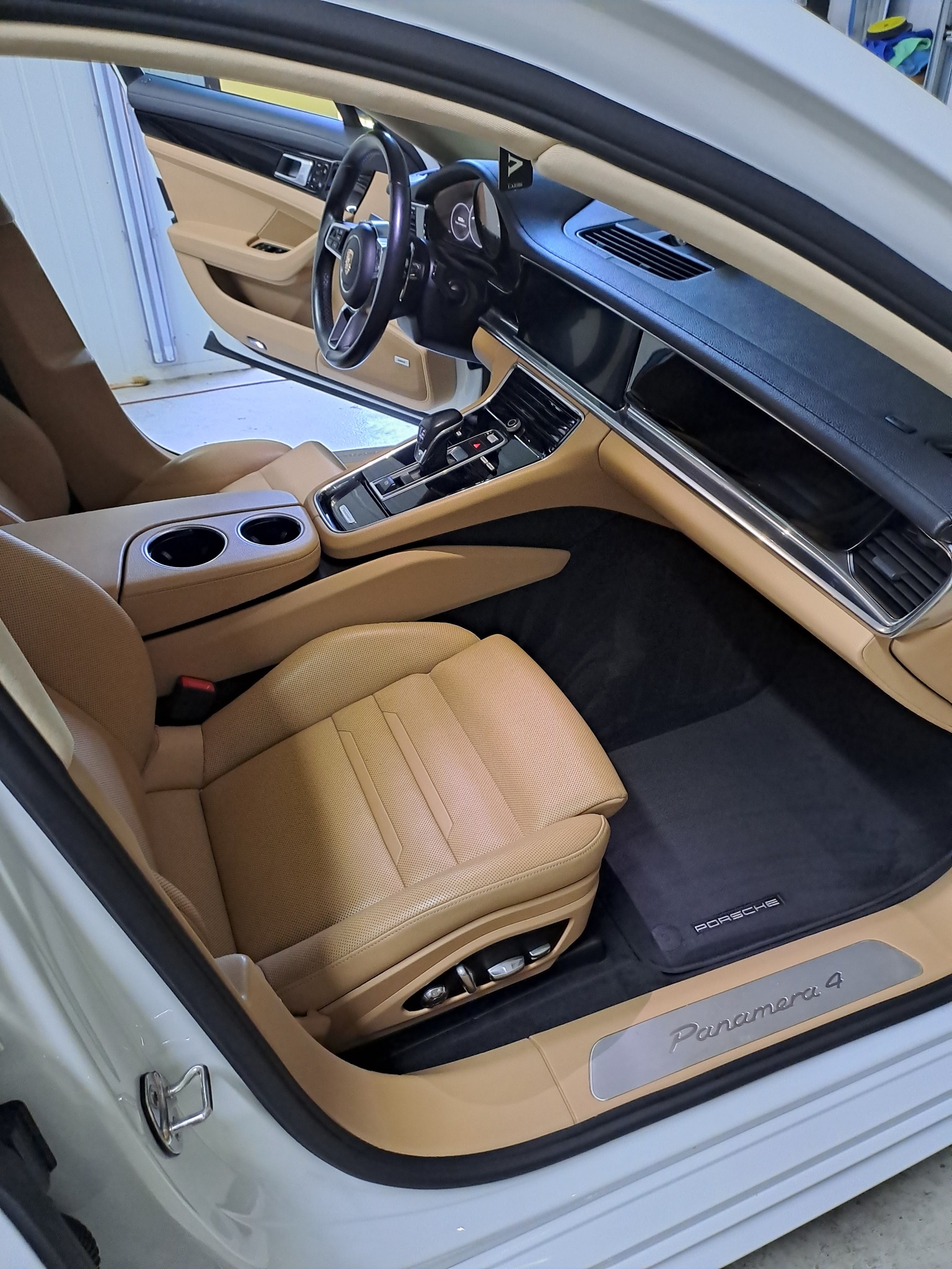 Wnętrze samochodu Porsche Panamera 4 z jasną skórzaną tapicerką, czarnymi dywanikami z logo marki, widok z otwartymi drzwiami.