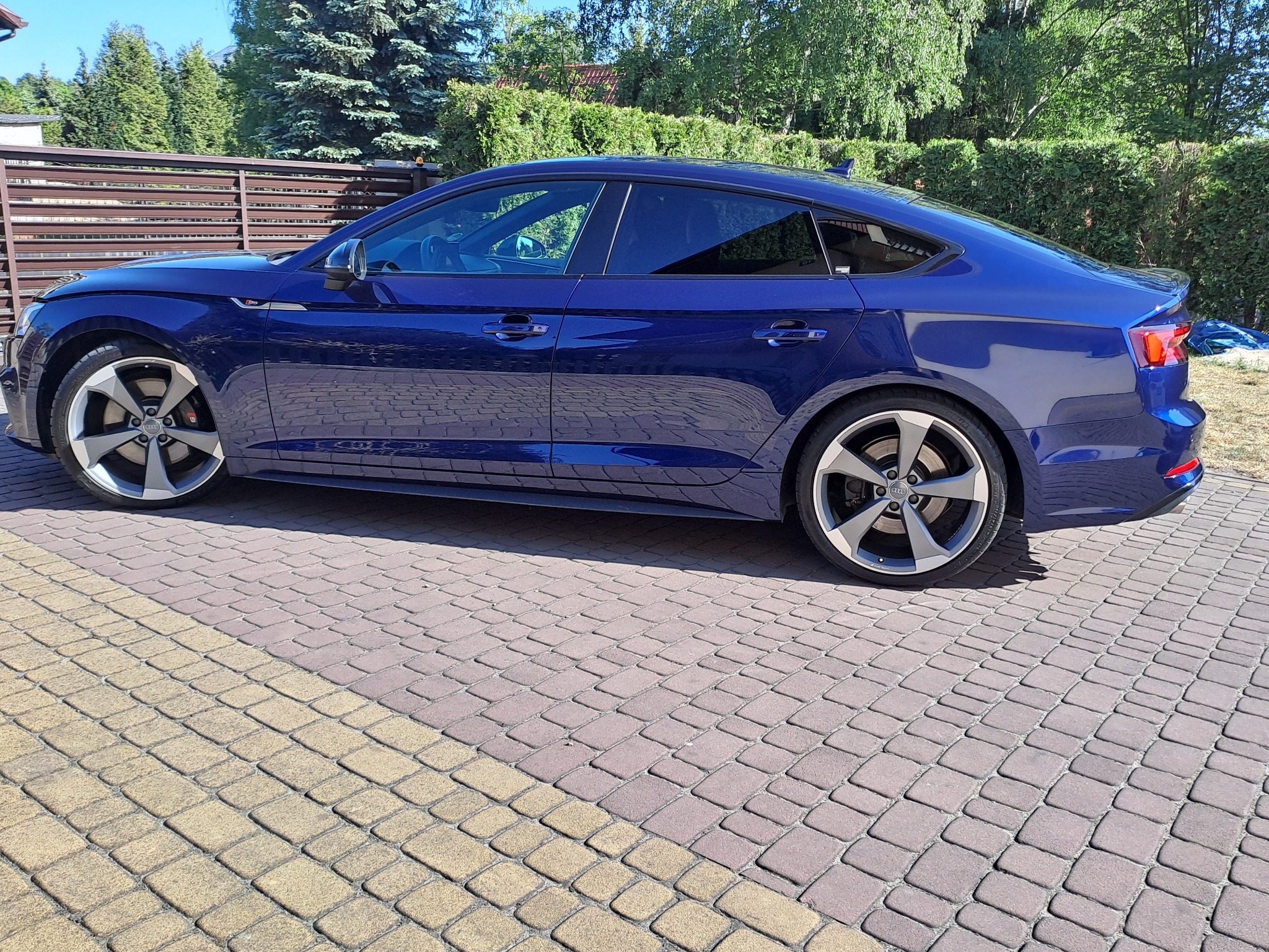 Niebieski samochód typu liftback z przyciemnionymi szybami bocznymi, zaparkowany na kostce brukowej w słoneczny dzień.