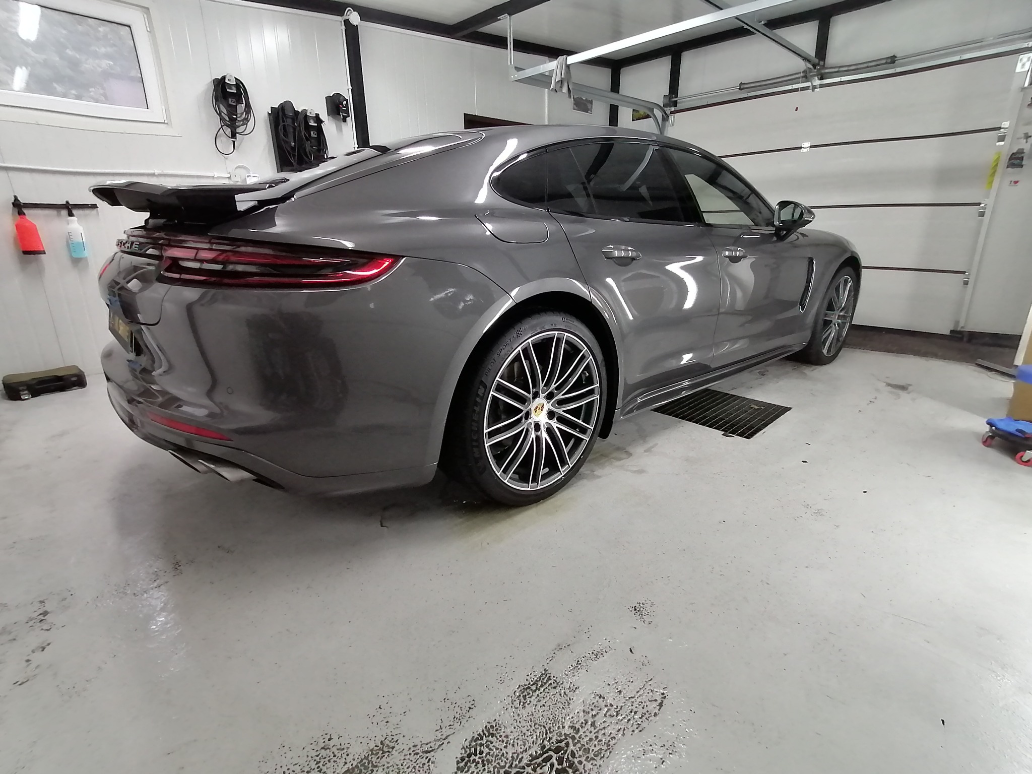 Szary samochód Porsche Panamera po umyciu, widoczne krople wody na podłodze garażu, felgi aluminiowe z czarnymi i srebrnymi ramionami.
