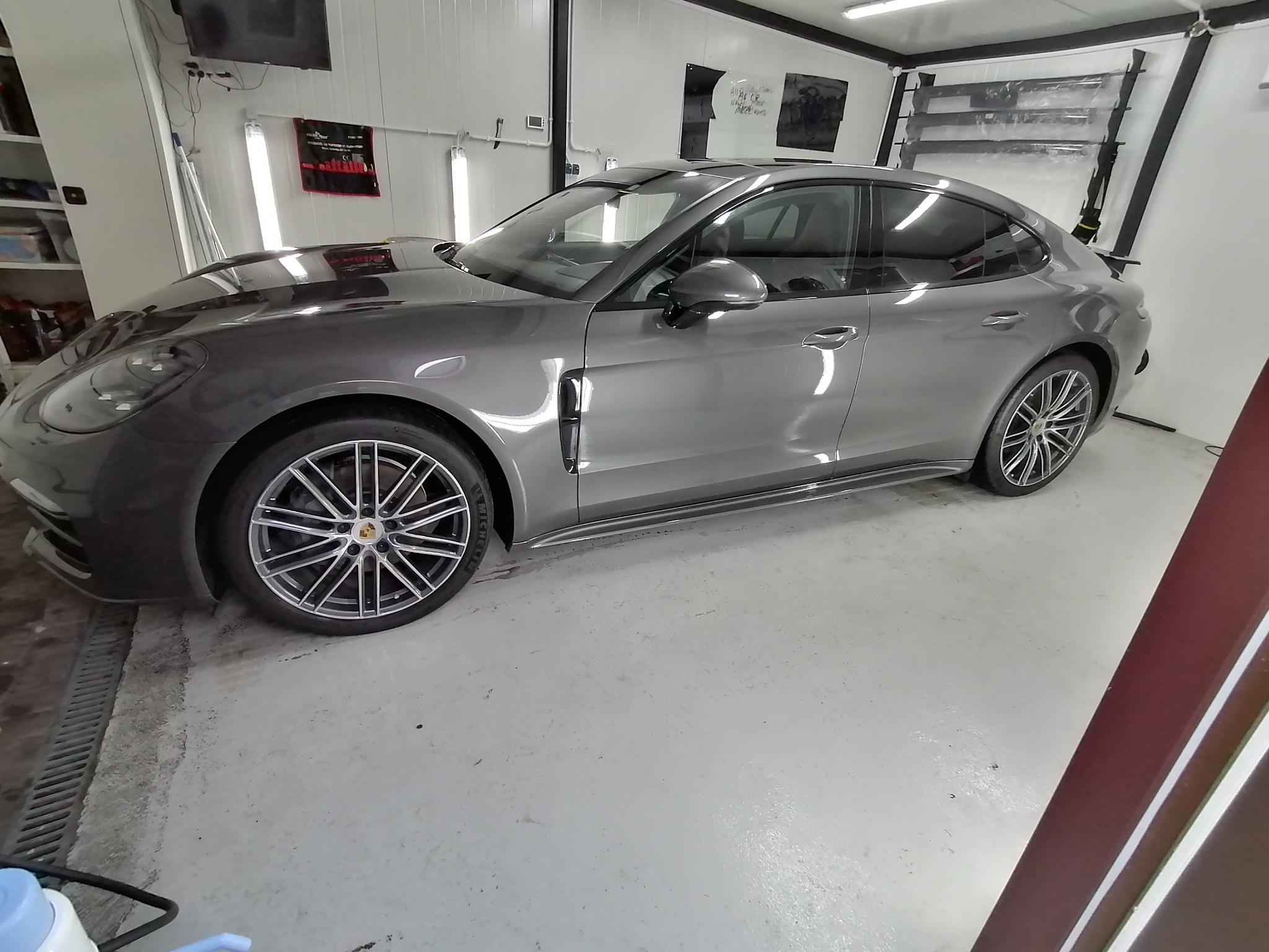 Szary samochód Porsche Panamera w sterylnym, jasnym garażu z foliami do oklejania szyb w tle, widoczne detale felg i karoserii.