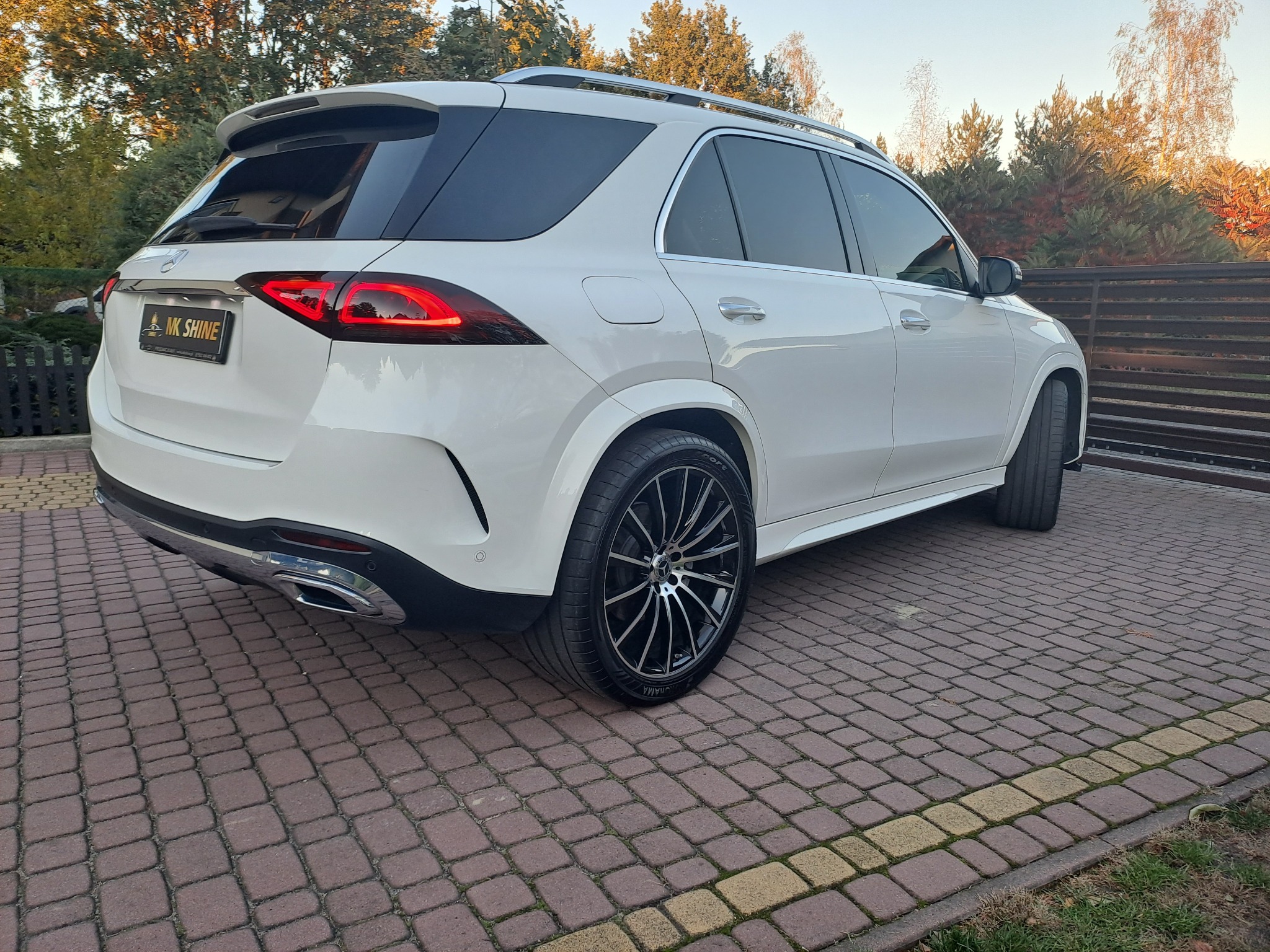 Biały SUV marki Mercedes z przyciemnionymi szybami, zaparkowany na kostce brukowej, z widocznym logo firmy MK Shine na tablicy rejestracyjnej.