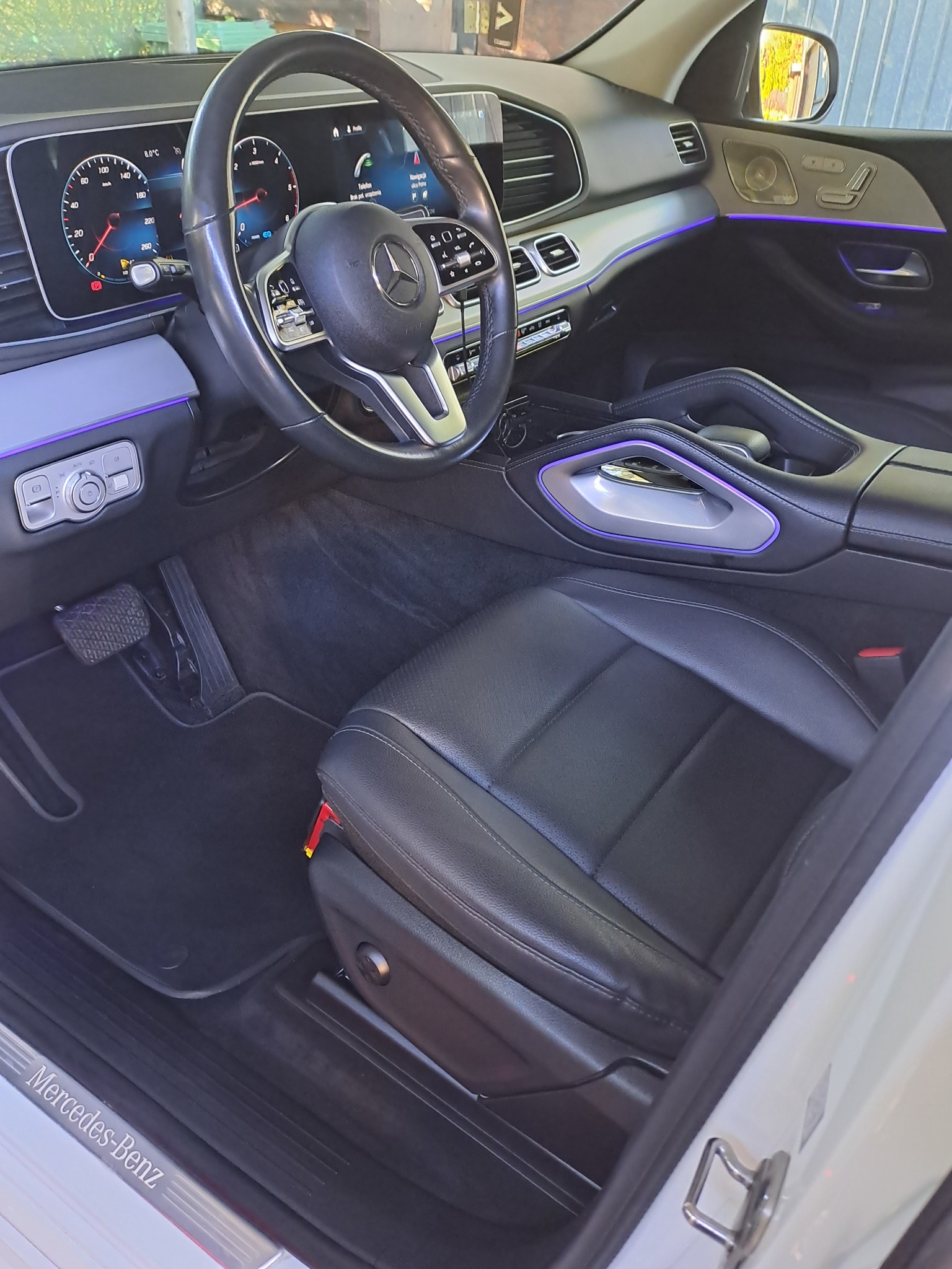 Wnętrze nowoczesnego samochodu marki Mercedes-Benz z czarną skórzaną tapicerką, podświetleniem LED w kolorze fioletowym i widocznym panelem sterowania.