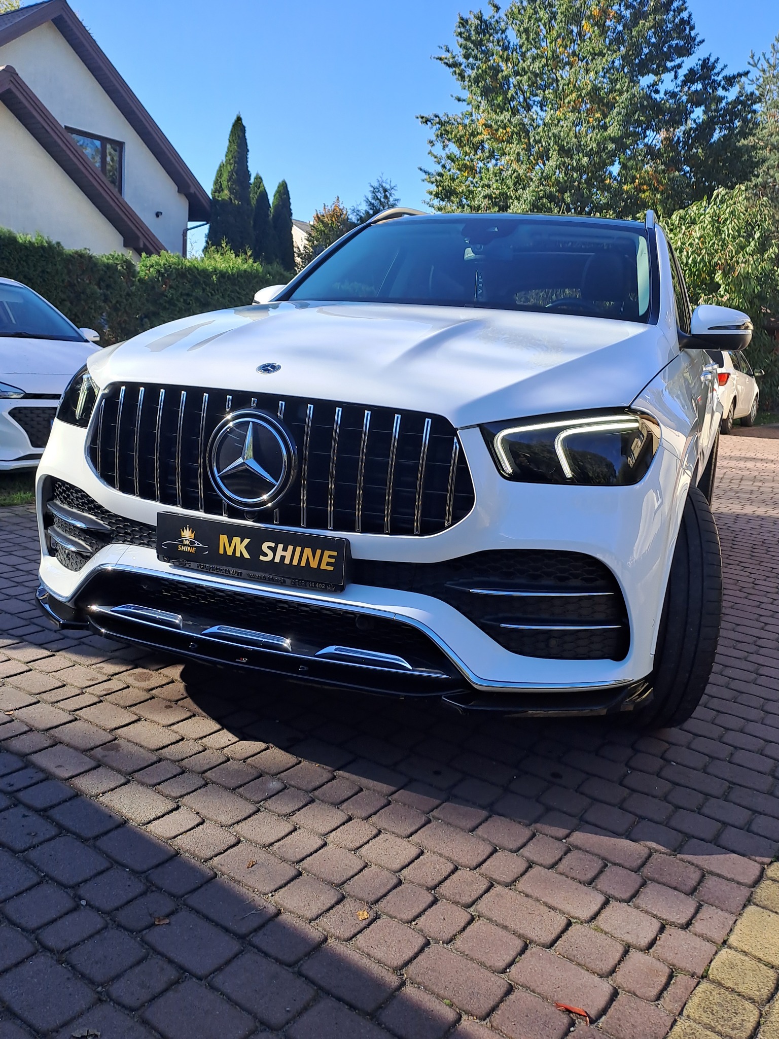 Biały Mercedes GLE po myciu, z widocznym logo MK Shine na przedniej tablicy rejestracyjnej, zaparkowany na kostce brukowej w słoneczny dzień.