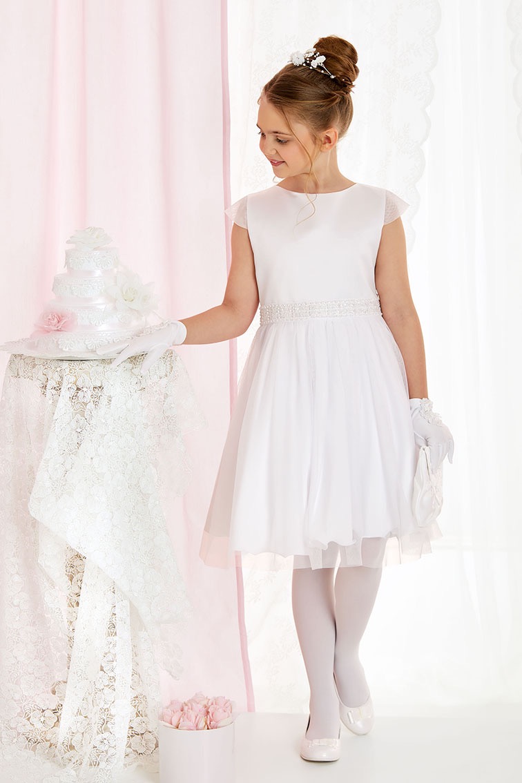 Dziewczynka w białej sukience komunijnej, z upiętymi włosami i ozdobą, pozuje obok tortu na stole przykrytym koronkowym obrusem, na tle jasnoróżowej zasłony.