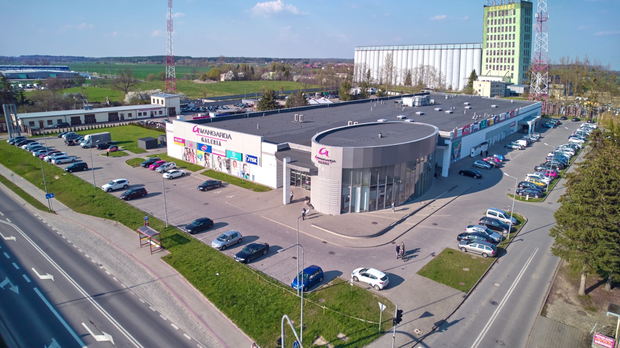 Zdjęcie z lotu ptaka przedstawia fasadę galerii handlowej Awangarda z parkingiem, otoczoną zielenią i drogami, w tle widoczny silos i wieża telekomunikacyjna.