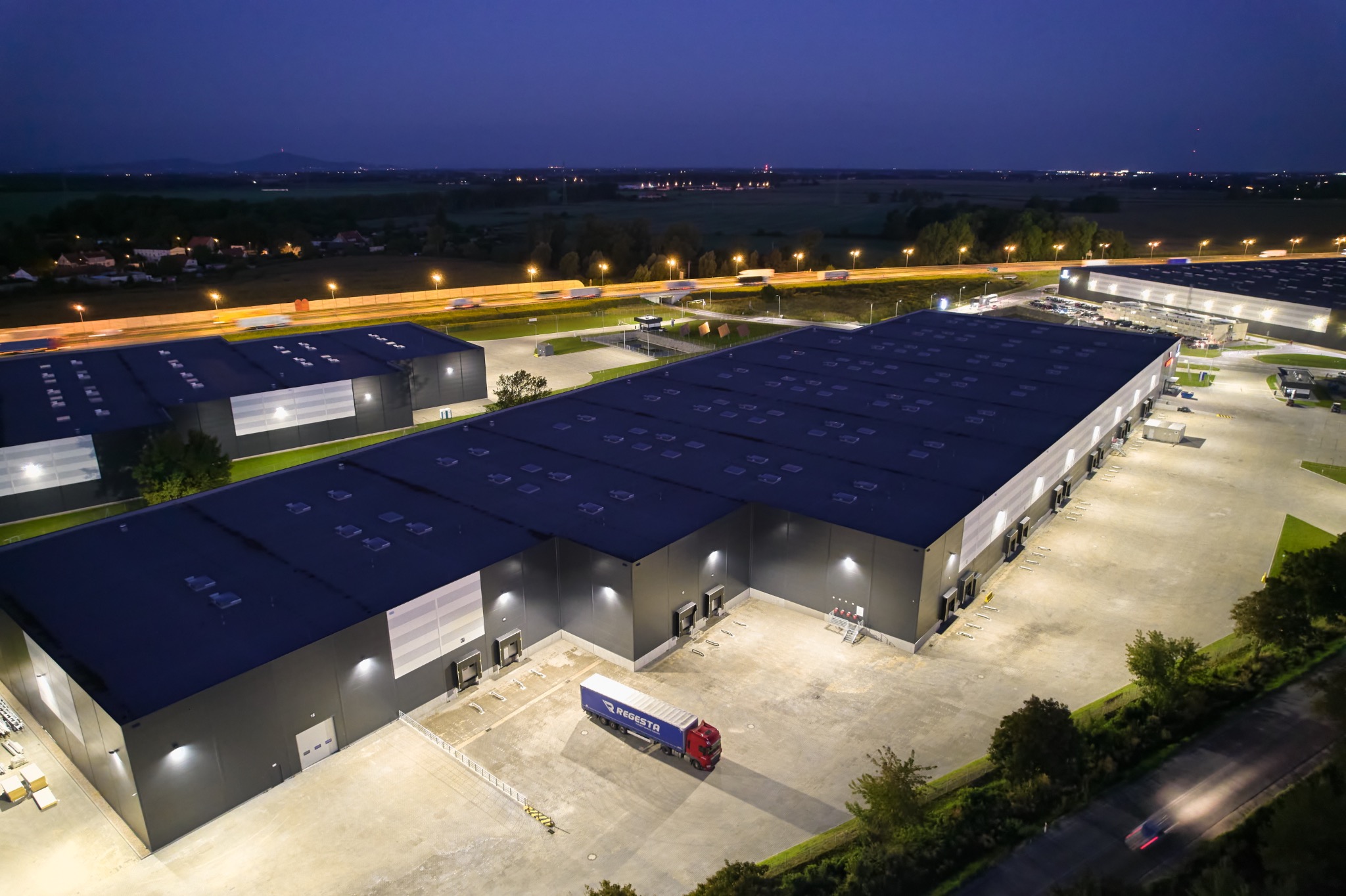 Nocny widok z lotu ptaka na kompleks magazynowy z zaparkowaną ciężarówką REGESSTA przed rampami załadunkowymi, w tle oświetlona autostrada i oddalone miasto.