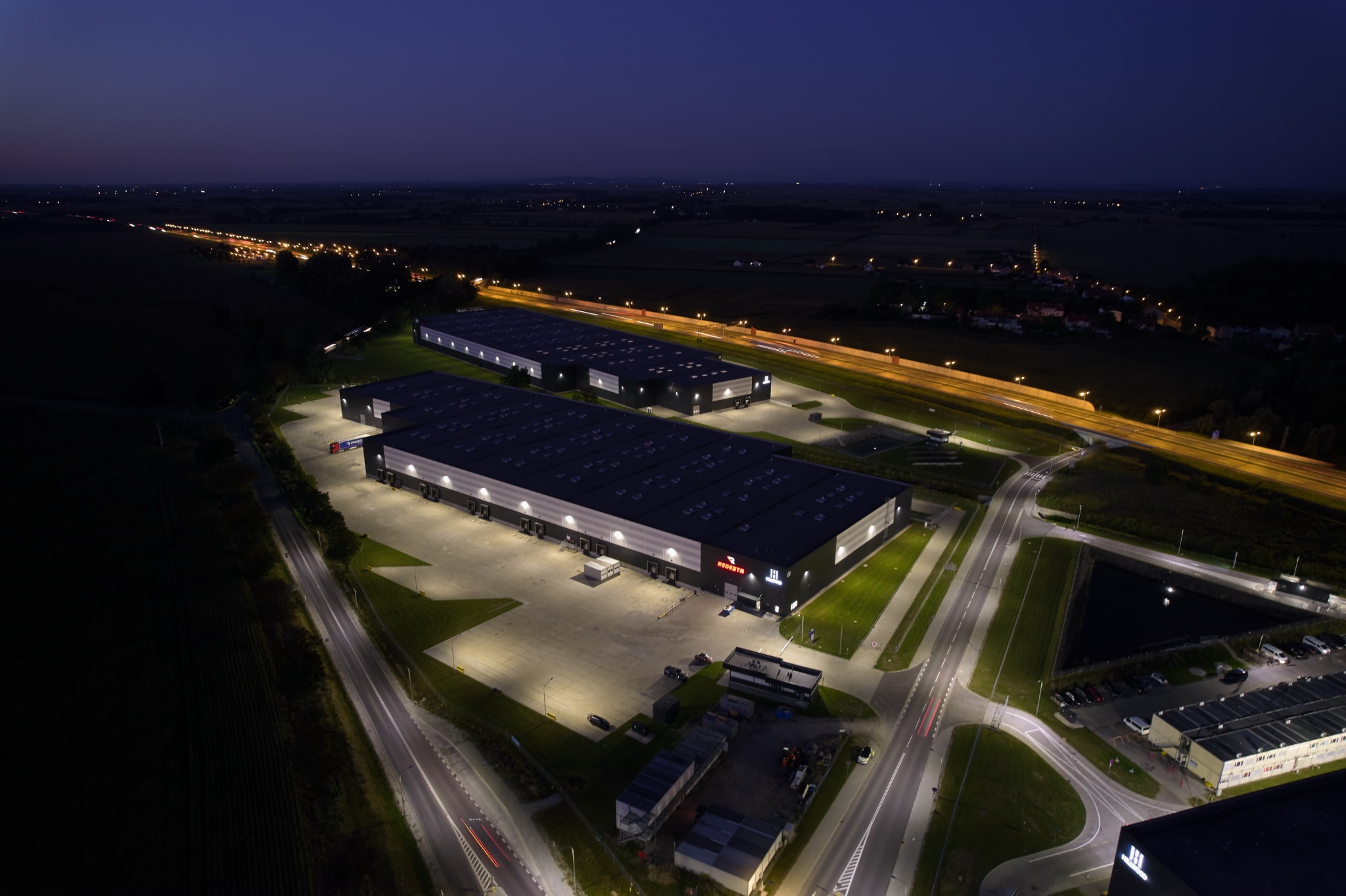Nocny widok z lotu ptaka na dwa oświetlone budynki magazynowe z logo firmy, położone obok autostrady z widocznymi smugami świateł samochodów.