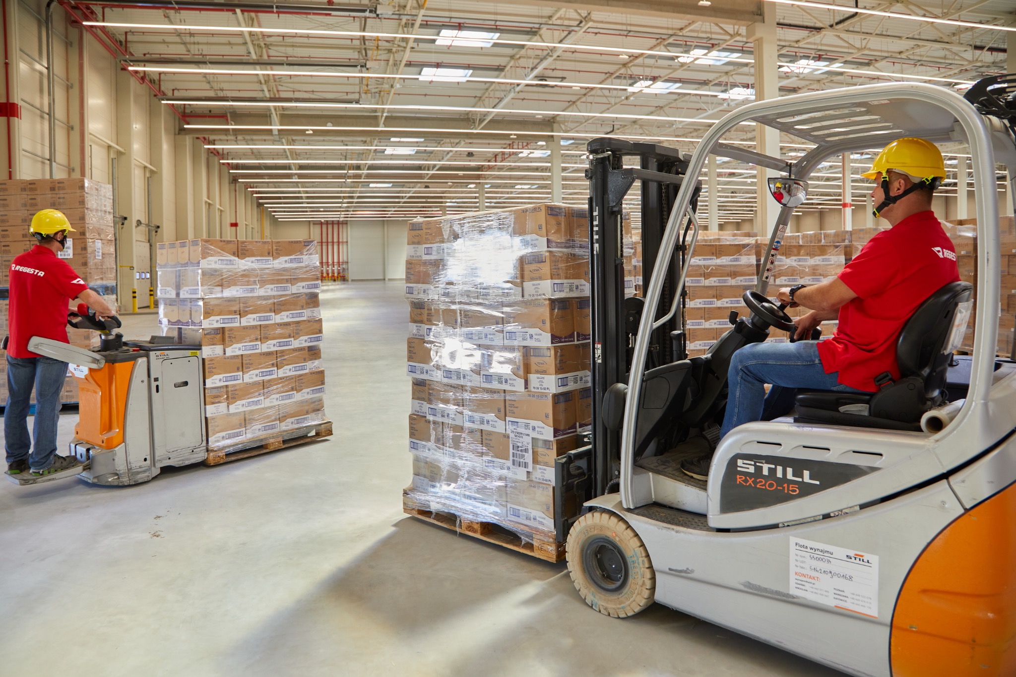 Wnętrze magazynu z dwoma operatorami wózków widłowych transportującymi palety z zapakowanymi kartonami. Jeden operator siedzi na wózku STILL RX20-15, drugi stoi obok wózka paletowego.