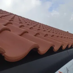 Czerwona dachówka ceramiczna ułożona rzędami, widok z dołu na tle pochmurnego nieba. Widoczny fragment rynny i folii z logo producenta dachówki.