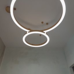 P&m bud - Nowoczesne oświetlenie sufitowe: dwa okrągłe, złote klosze LED zawieszone na cienkich linkach, oświetlające gładką, jasną ścianę z delikatnymi smugami.