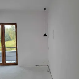 Pusty pokój w nowym budownictwie z białymi ścianami, szarą podłogą, drewnianymi drzwiami balkonowymi i minimalistyczną czarną lampą wiszącą.