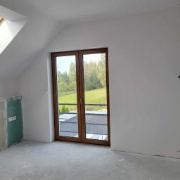 P&m bud - Poddasze w trakcie wykańczania: ściany pomalowane na biało, drewniane okno balkonowe z widokiem na zieleń, fragment ściany z płyt kartonowo-gipsowych przed wykończeniem, surowa wylewka na podłodze.