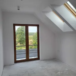 Poddasze po szpachlowaniu i malowaniu na biało, z drewnianymi oknami dachowymi i balkonowymi, surowa betonowa podłoga przed dalszym wykończeniem.