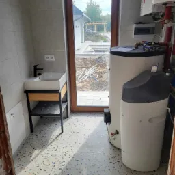 Wnętrze pomieszczenia gospodarczego z dwoma zbiornikami na wodę, umywalką na metalowym stelażu z drewnianymi elementami oraz widokiem na plac budowy przez okno.