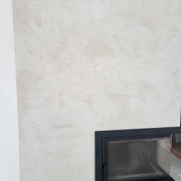 P&m bud - Ściana z dekoracyjną, jasnoszarą fakturą przypominającą beton, z wkomponowanym czarnym wkładem kominkowym i poziomym czarnym panelem wentylacyjnym u góry.