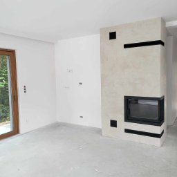 P&m bud - Surowe wnętrze z wykończonym kominkiem o kremowej, teksturowanej powierzchni i czarnych akcentach, na tle białych ścian i surowej posadzki, widoczne gniazdka elektryczne i okno z widokiem na zieleń.