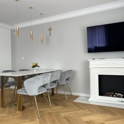 P&m bud - Jasne, nowocześnie urządzone wnętrze jadalni z parkietem ułożonym w jodełkę, marmurowym stołem, szarymi krzesłami na złotych nogach, dekoracyjnym kominkiem i telewizorem na ścianie, złote lampy...