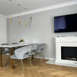 Jasne, nowocześnie urządzone wnętrze jadalni z parkietem ułożonym w jodełkę, marmurowym stołem, szarymi krzesłami na złotych nogach, dekoracyjnym kominkiem i telewizorem na ścianie, złote lampy...