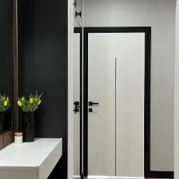 Nowoczesny przedpokój z białymi drzwiami z czarną ramą, lustrem, konsolą z żółtymi tulipanami w czarnym wazonie i drewnianą podłogą.