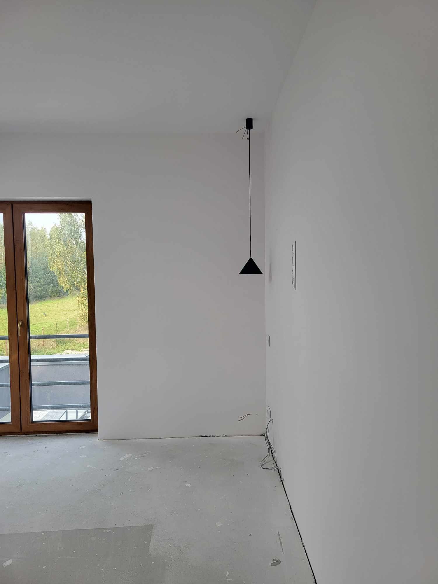 Pusty pokój w nowym budownictwie z białymi ścianami, szarą podłogą, drewnianymi drzwiami balkonowymi i minimalistyczną czarną lampą wiszącą.
