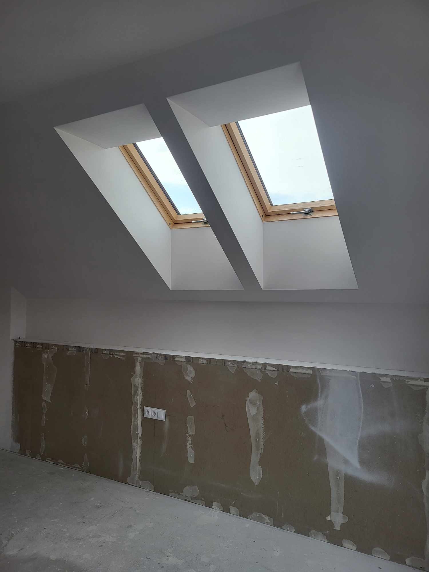 Poddasze w trakcie remontu z dwoma oknami dachowymi; ściany przygotowane do dalszych prac, widoczne ślady po usuniętych elementach i gniazdko elektryczne.