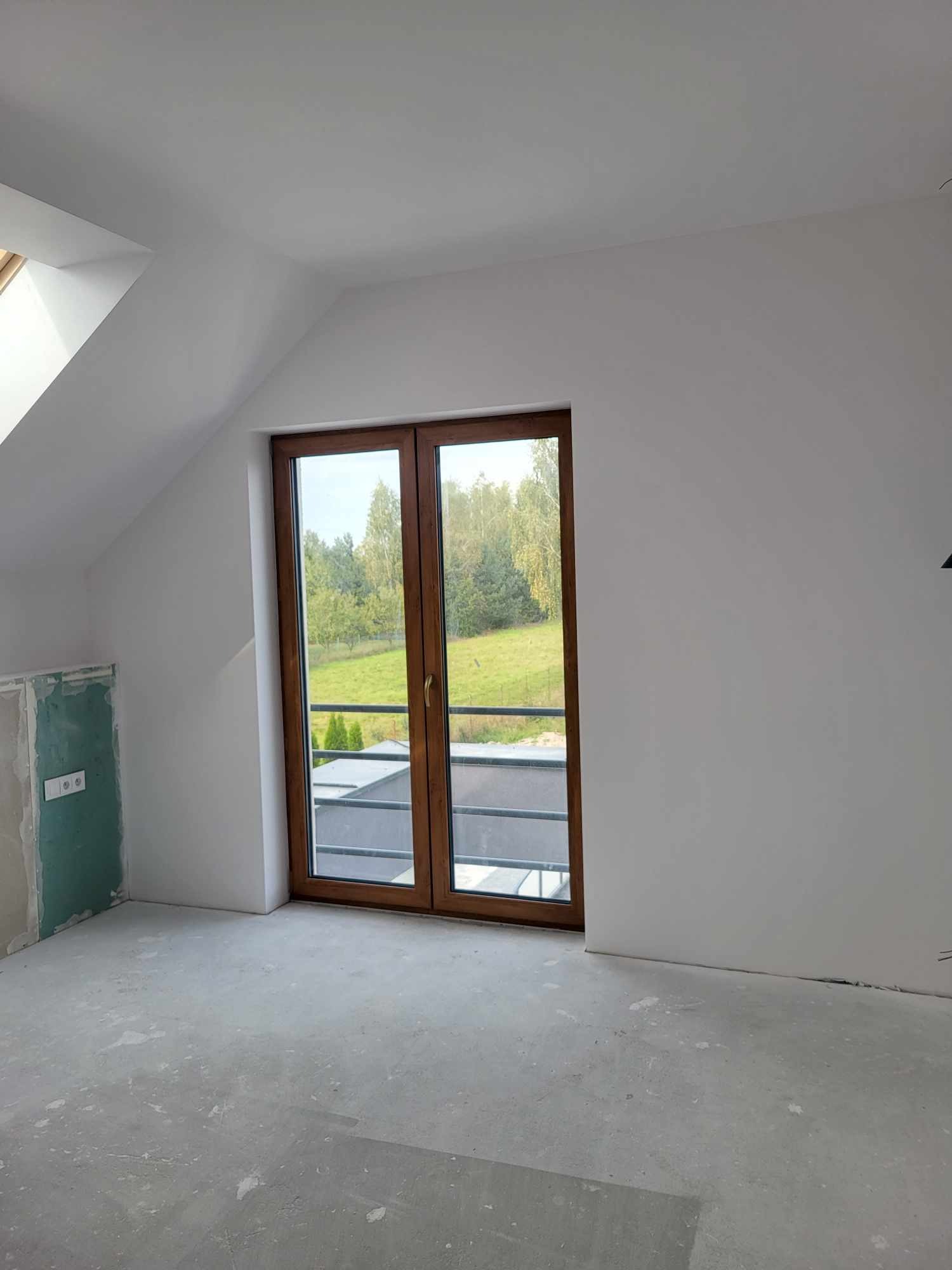 Poddasze w trakcie wykańczania: ściany pomalowane na biało, drewniane okno balkonowe z widokiem na zieleń, fragment ściany z płyt kartonowo-gipsowych przed wykończeniem, surowa wylewka na podłodze.
