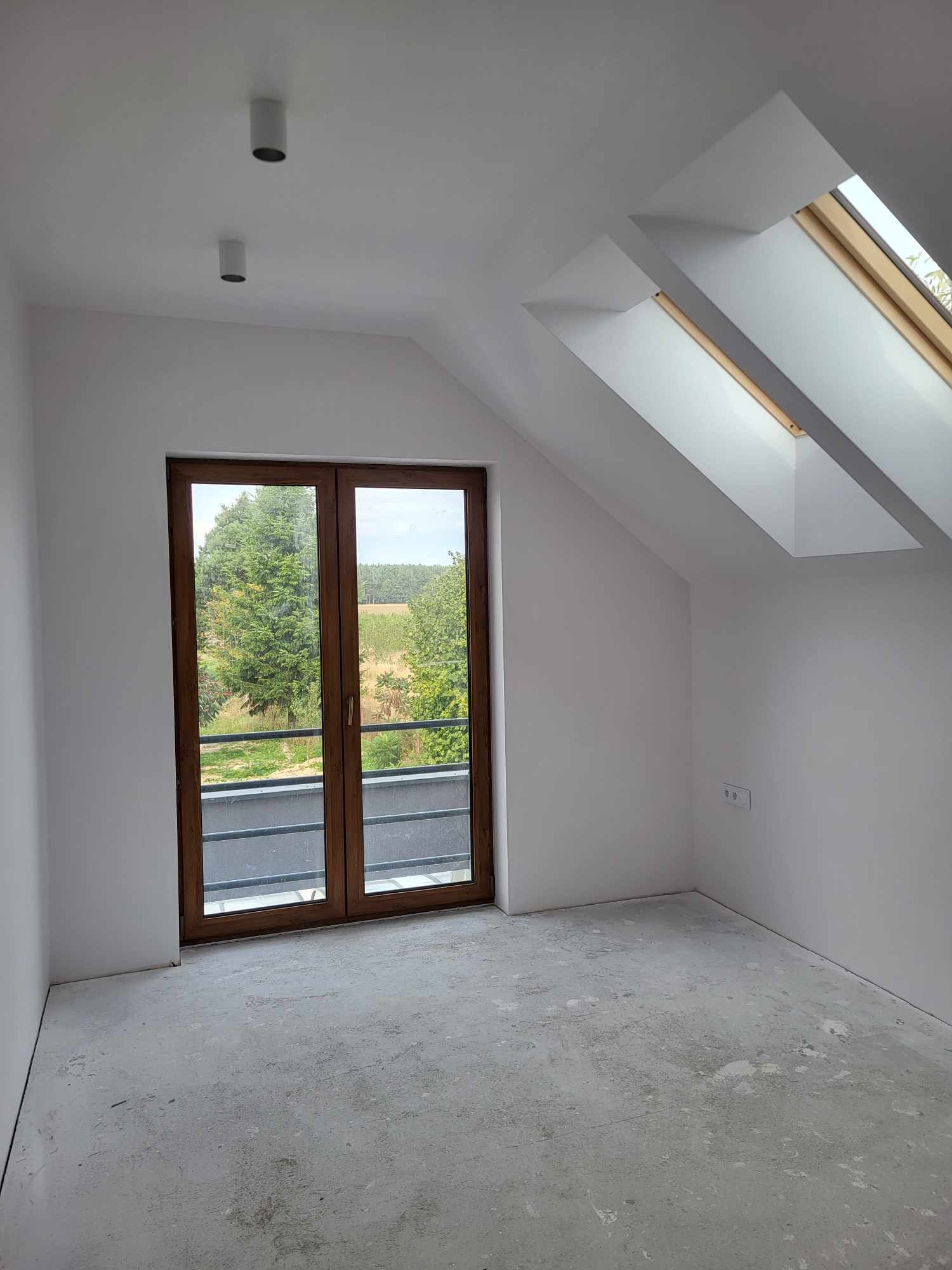 Poddasze po szpachlowaniu i malowaniu na biało, z drewnianymi oknami dachowymi i balkonowymi, surowa betonowa podłoga przed dalszym wykończeniem.