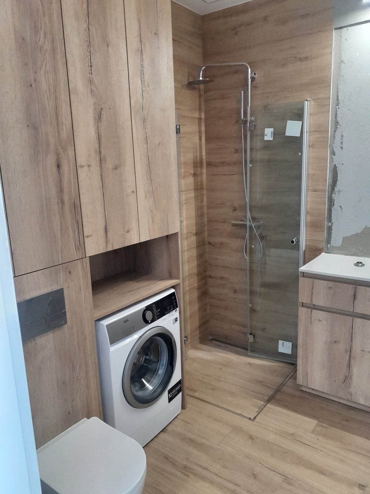 Nowoczesna łazienka z drewnopodobnymi płytkami, kabiną prysznicową typu walk-in, zabudową meblową z pralką AEG i podwieszaną toaletą.