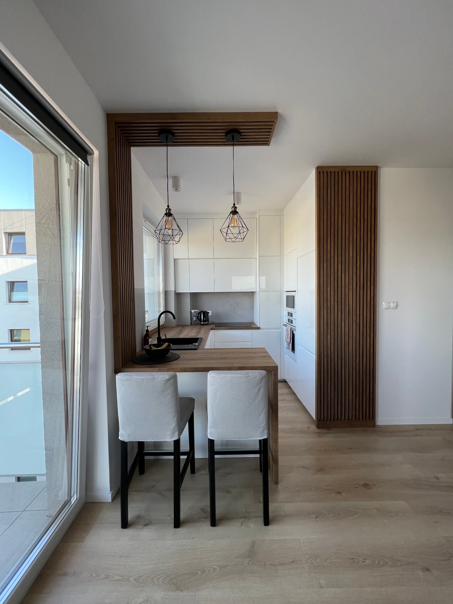 Nowoczesna kuchnia z białymi szafkami, drewnianym blatem, dekoracyjnymi lamelami ściennymi i dwoma krzesłami barowymi z jasnymi pokrowcami, widziana z perspektywy balkonu.