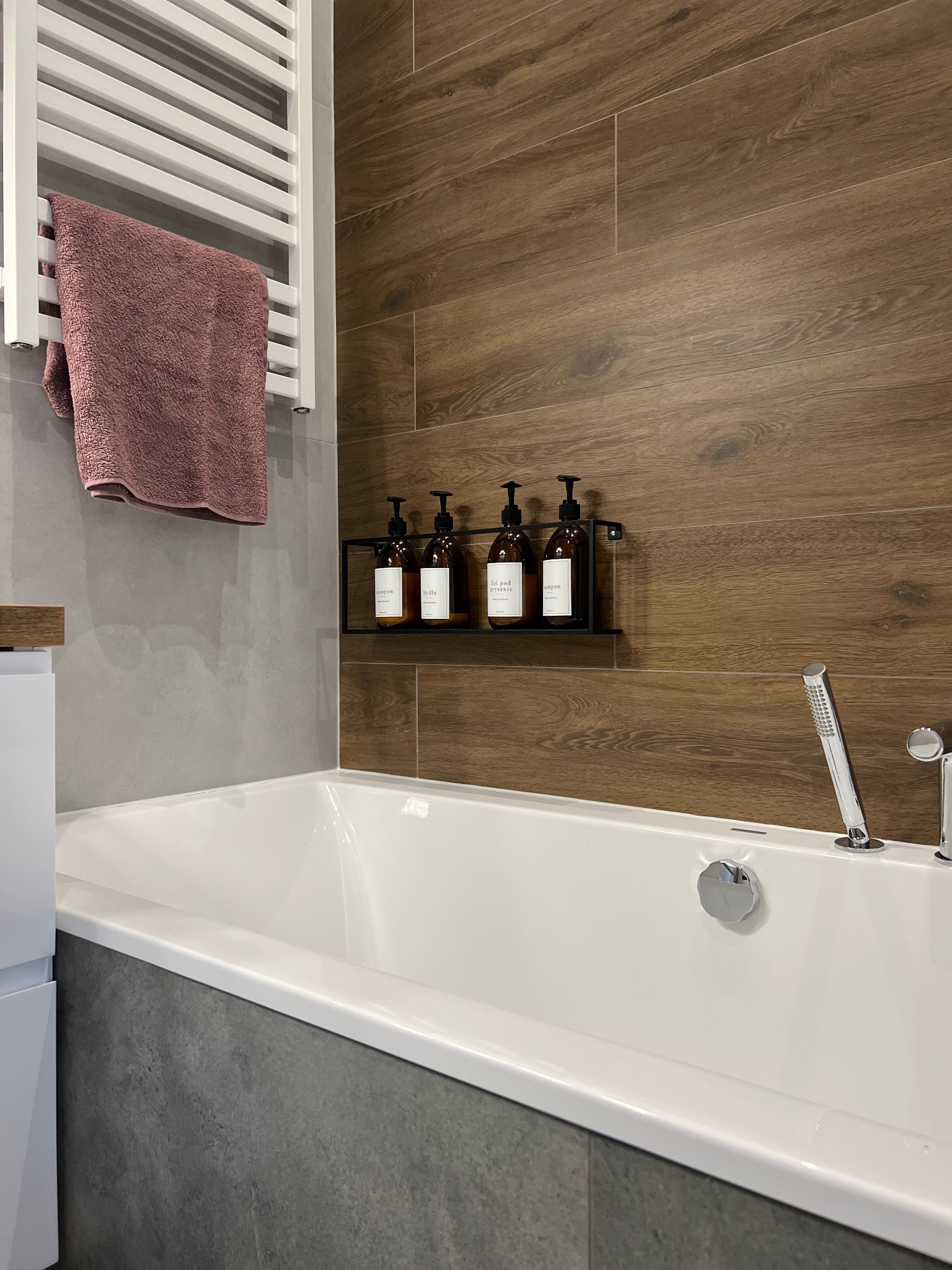 Nowoczesna łazienka z wanną, brązowymi płytkami imitującymi drewno, szarą ścianą, grzejnikiem drabinkowym z różowym ręcznikiem i zestawem brązowych butelek na kosmetyki.