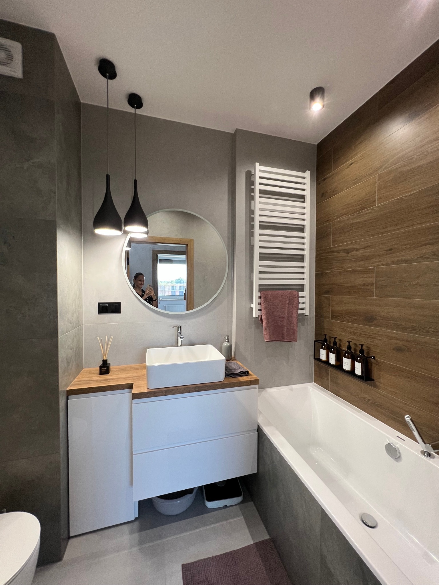 Nowoczesna łazienka z drewnianymi i szarymi płytkami, białym grzejnikiem drabinkowym z powieszonym ręcznikiem, wiszącymi czarnymi lampami nad umywalką nablatową i okrągłym lustrem, w którym odbija...