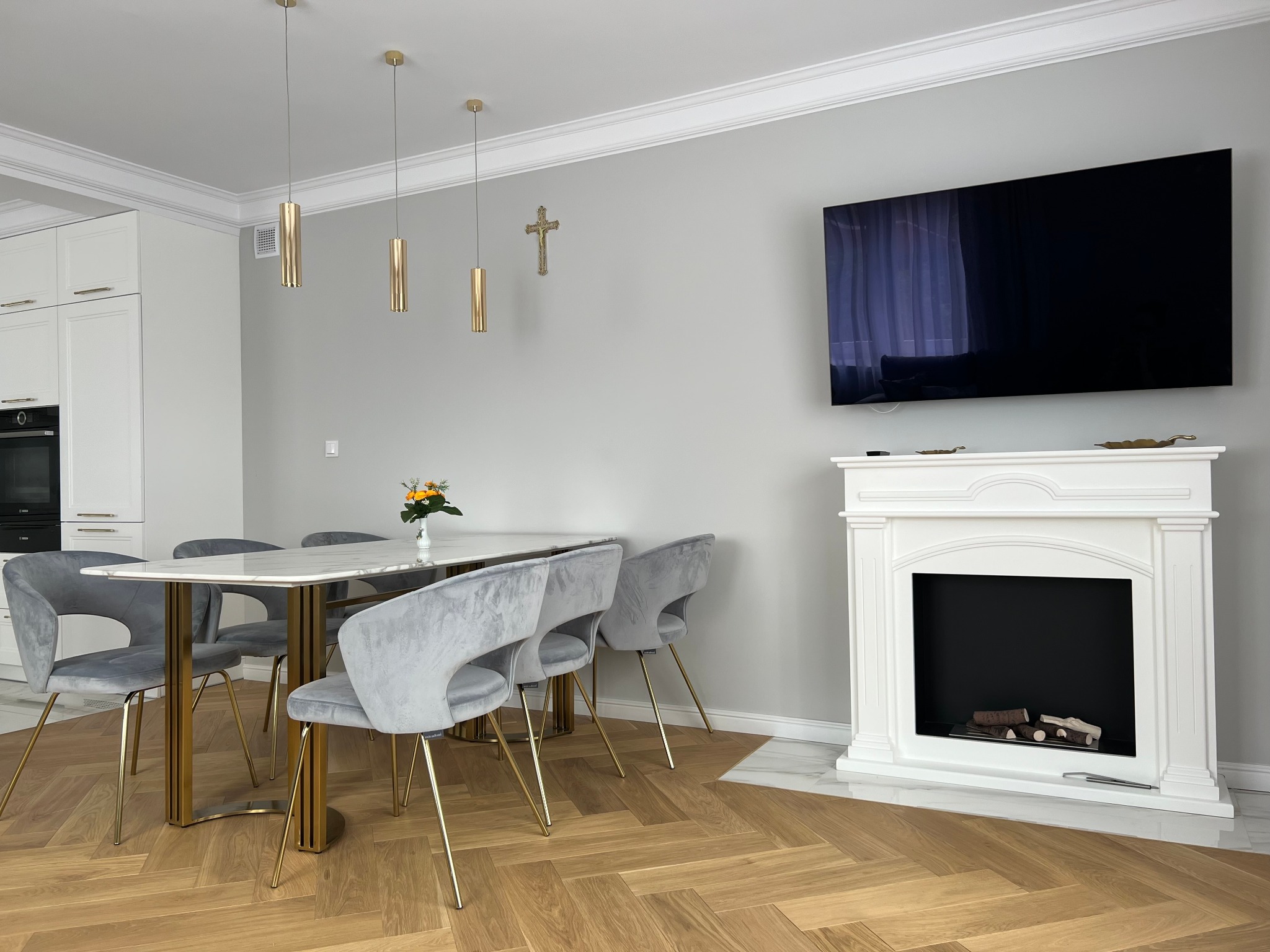 Jasne, nowocześnie urządzone wnętrze jadalni z parkietem ułożonym w jodełkę, marmurowym stołem, szarymi krzesłami na złotych nogach, dekoracyjnym kominkiem i telewizorem na ścianie, złote lampy...