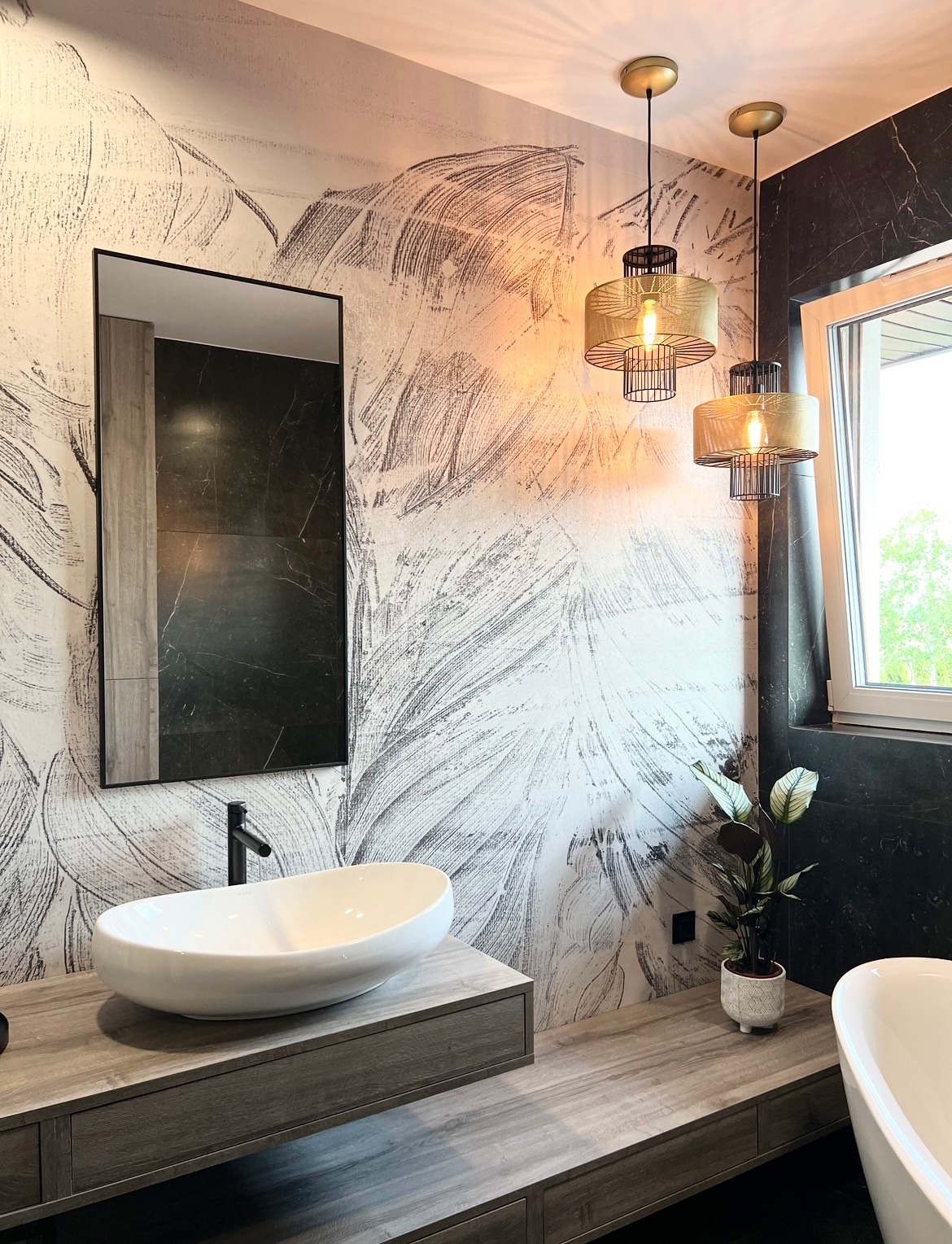 Nowoczesna łazienka z umywalką nablatową o owalnym kształcie, czarną armaturą i dużym lustrem w czarnej ramie na tle ściany z dekoracyjnym wzorem przypominającym liście, oświetlona dwoma wiszącymi...